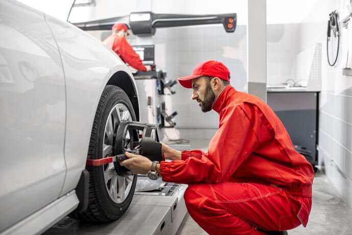 Imagem de um homem agachado com um macacão de mecânica e um boné vermelho. Ele está de frente para uma roda de um carro, utilizando uma ferramenta de alinhamento de pneu.